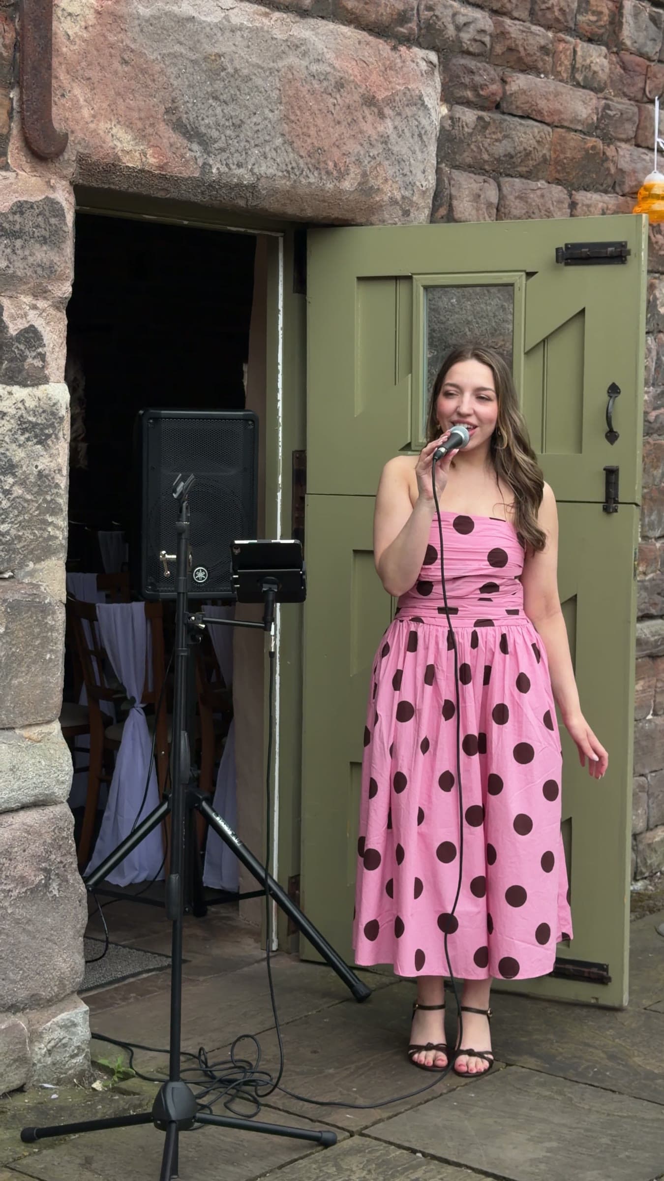 Olivia singing at a drinks reception in a stone courtyard, in a pink polka-dot dress