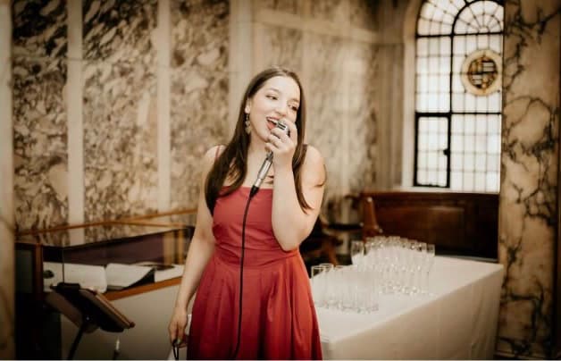 Olivia singing in a red dress in a marble hall, with champagne glasses set out